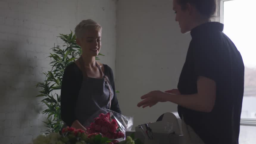 A woman working at a flower shop gestures toward the flowers on a counter that another woman is buying