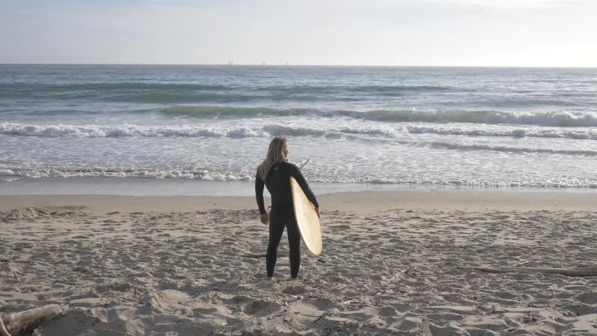 A woman wearing a wetsuit looks around at the ocean while holding a surfboard on a beach