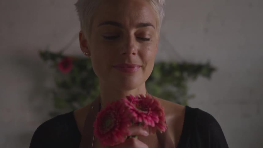 A woman sniffs a flower held in her hand then turns her head to the left indoors
