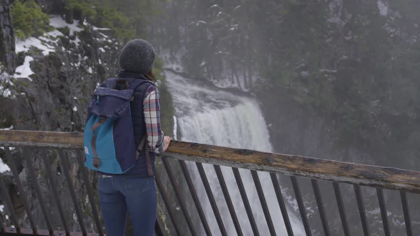 Woman wearing backpack looks over waterfall in winter