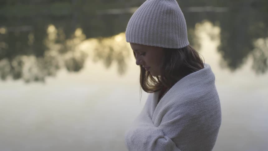 Female in a robe looks down while standing next to a body of water
