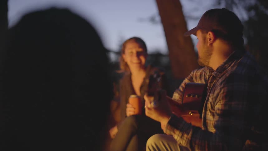 A man sitting by a fire with other people strums a guitar and sings