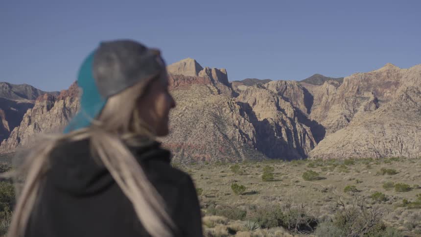 A woman wearing a hat with long blond hair sits and looks out at a beautiful mountain range on a bright sunny day