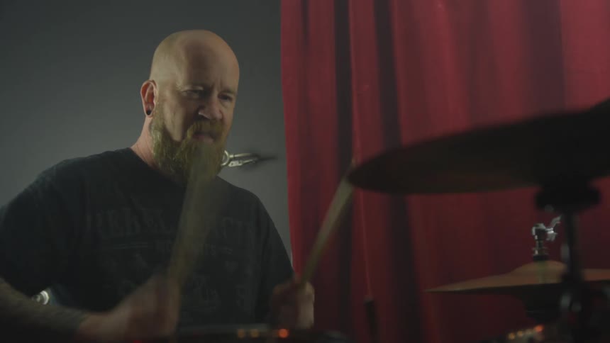 A bald, bearded man plays the drums