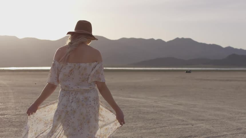 Woman in flowing dress walks across desert flats in front of mountains