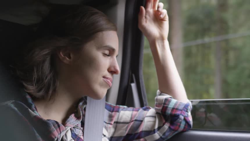 A young person allows the wind to pour in through the window of a car driving down the road and their hair waves in the wind wearing a seatbelt with their eyes closes on a bright sunny day