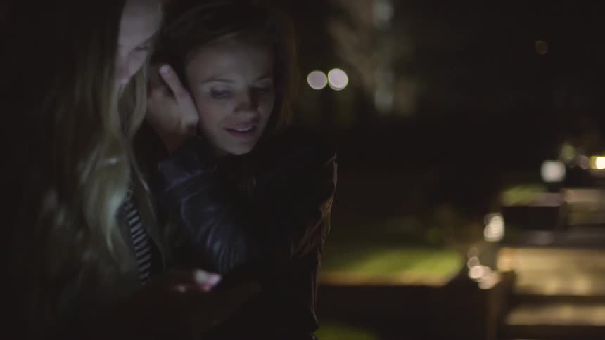Two women talk and laugh while looking at phone at night