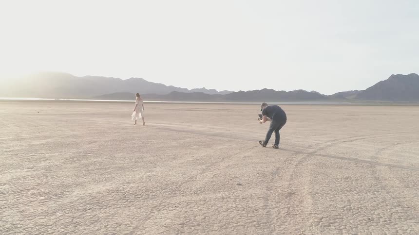Man films a woman as she walks away in the desert