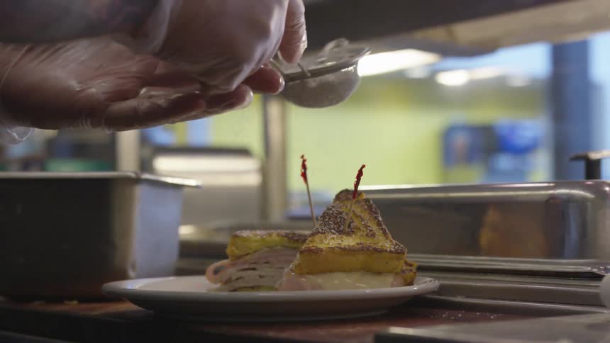A restaurant worker sprinkles an ingredient over a plated sandwich