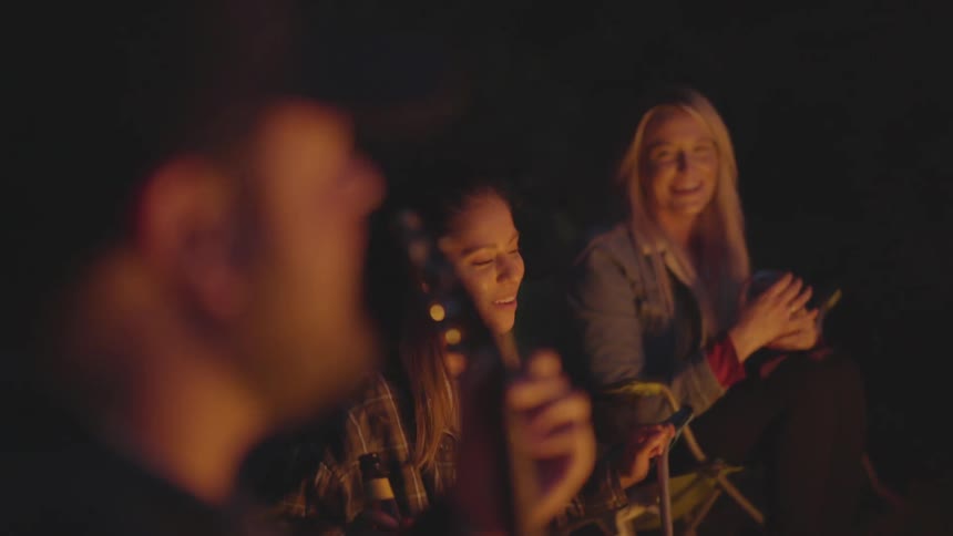 A man plays a guitar and sings next to a campfire with friends