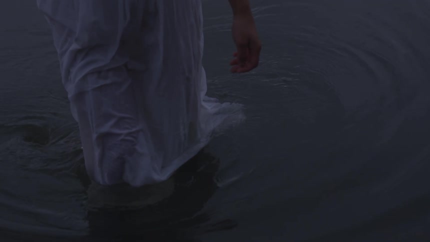 A woman in a soaked white dress walks out of the water