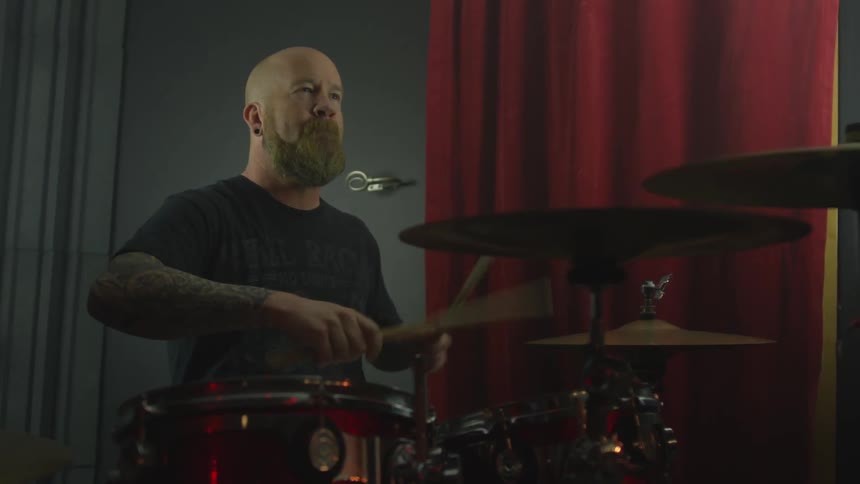 A man with a shaved head and a thick beard is playing the drums