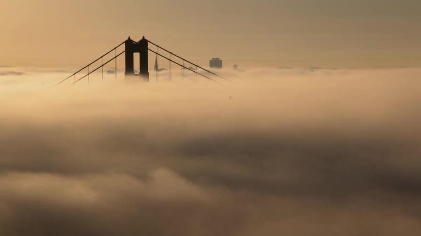 Clouds glide around a suspended bridge near a city