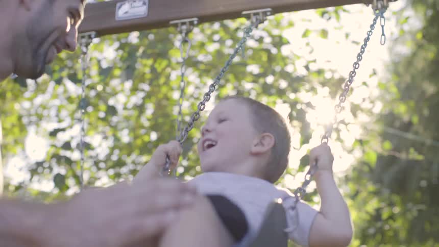 A young white boy is being pushed on a swing by his dad in a backyard with a privacy fence