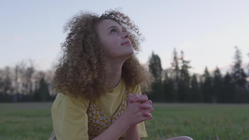 Girl sitting in a field with her hands cupped while staring into the sky