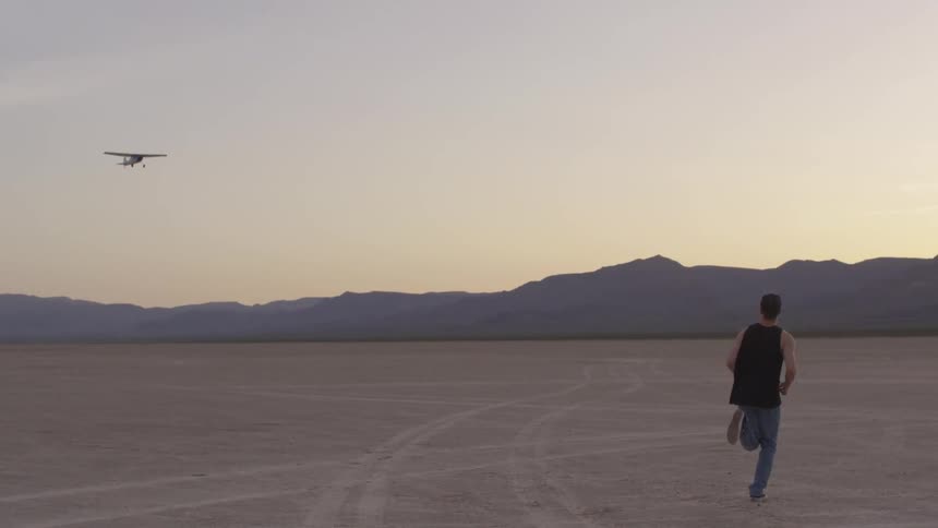 A man runs out to the dry barren cracking soil sands of a desert landscape as the sun sets and an airplane flies above him