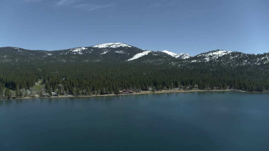 The camera zooms and flies towards the shoreline of a beautiful forest with snow capped mountains in the background on a bright sunny day