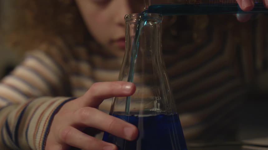 A little girl pours blue liquid into a narrow mouthed beaker