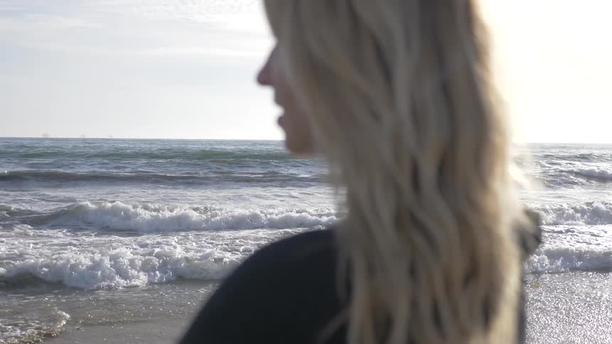 A woman with blond hair looks at the horizon as she stands facing an ocean