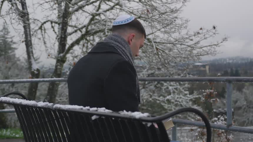 A man wearing a yamaka sits on a bench that has snow on it