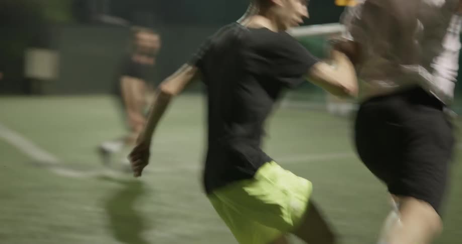 Men play soccer and step out of bounds on a dark night with a green grassy field lit up by bright lights