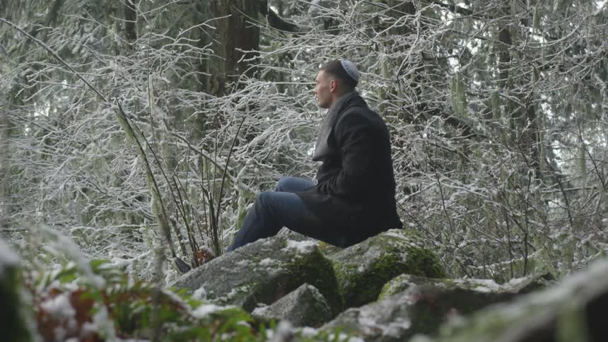 Young jewish man sits on large rocks in forest with light snow