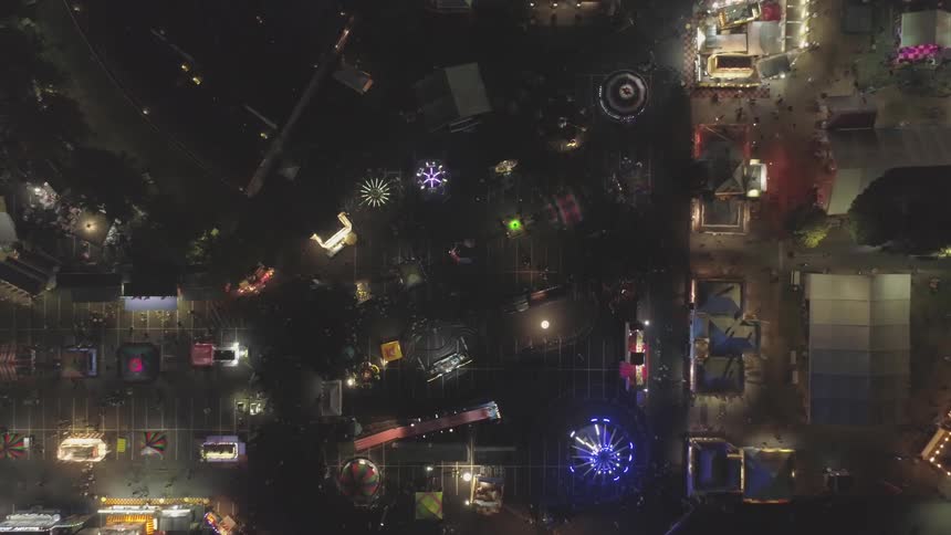 Camera zooms out overlooking a amusement park filled with neon lights with an aerial view on a dark night