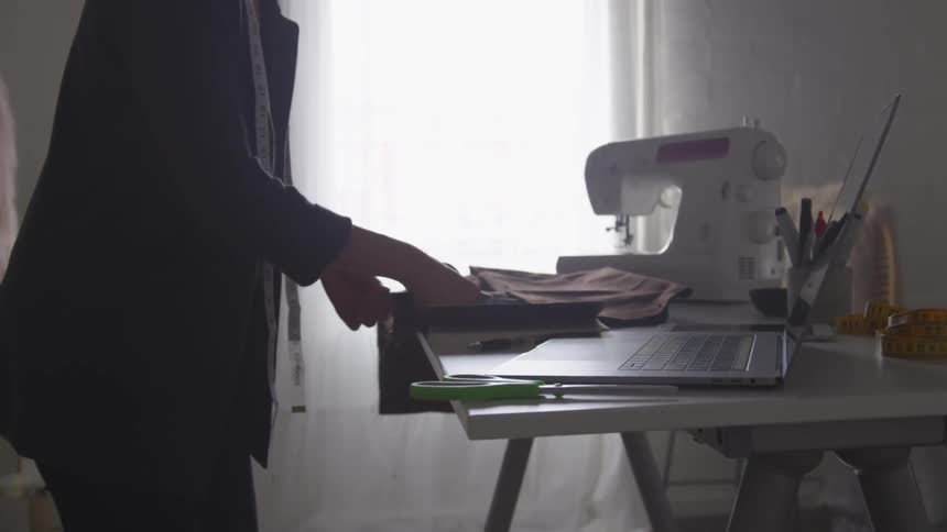 Woman standing at desk with computer and sewing machine cuts fabric with scissors
