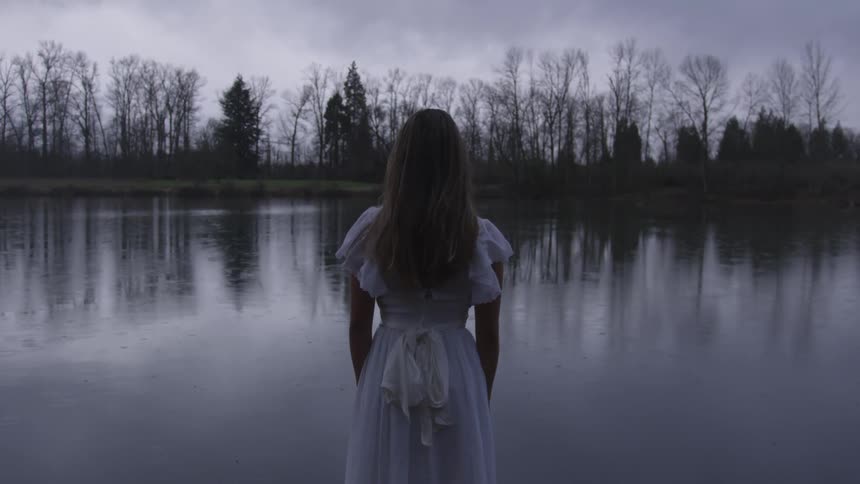 Young woman in white dress stands in front of lake