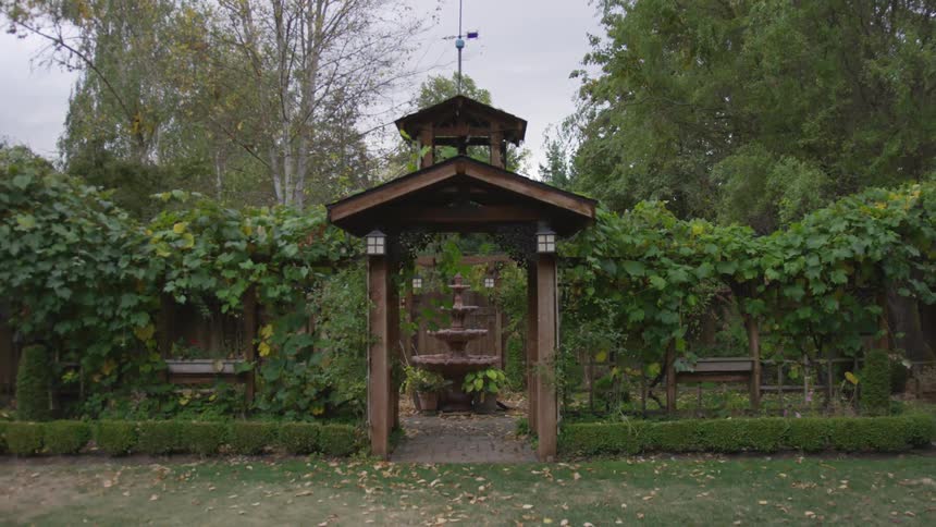 Garden arbor topped with weather vane leads into vine covered garden with water fountain