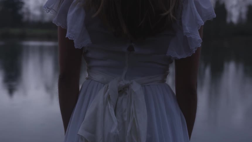 A girl in a white dress standing in front of a still lake at dusk