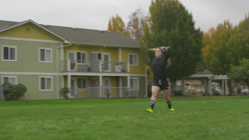 Woman in soccer apparel points and throws soccer ball over head to teammate in yard