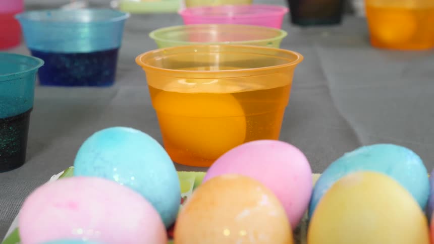Different colored easter eggs sit in a carton as more are being dyed in cups