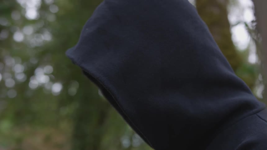 A man has his hood up covering his face as he stands in a forest