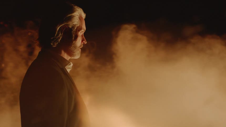 A man stands in a cloud of smoke blowing past him while he stands beside a fire