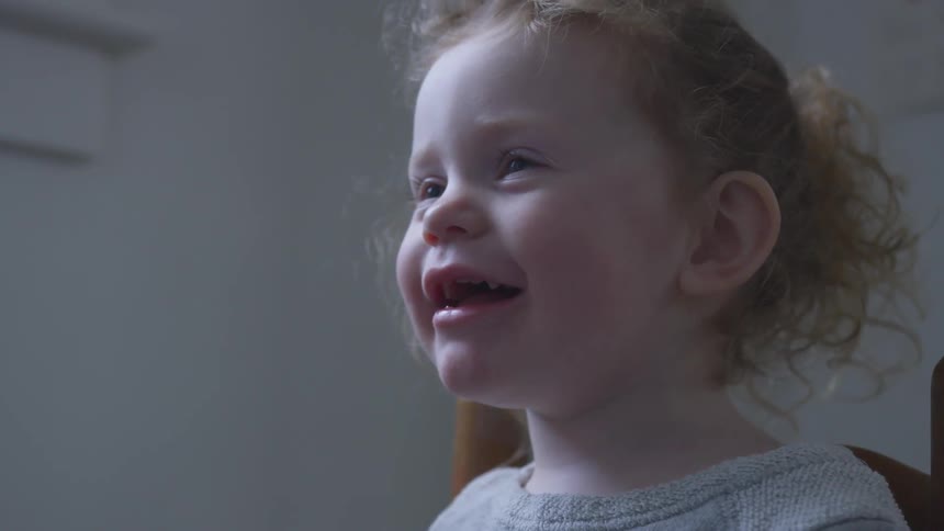 A little girl smiles and laughs as she looks up to talk at someone and then back down in a dim lit room