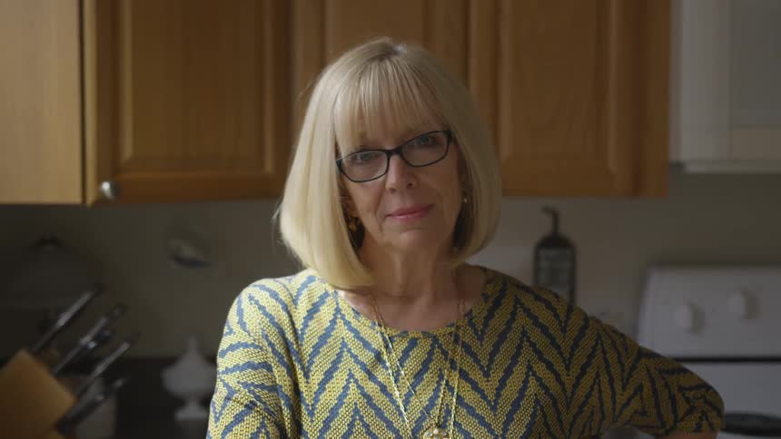 An elderly women looks out the window with blond hair and black rim glasses inside her home kitchen