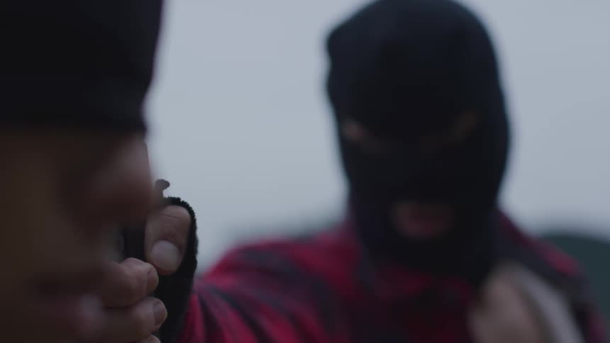 A person in a ski mask points a gun at a person blindfolded