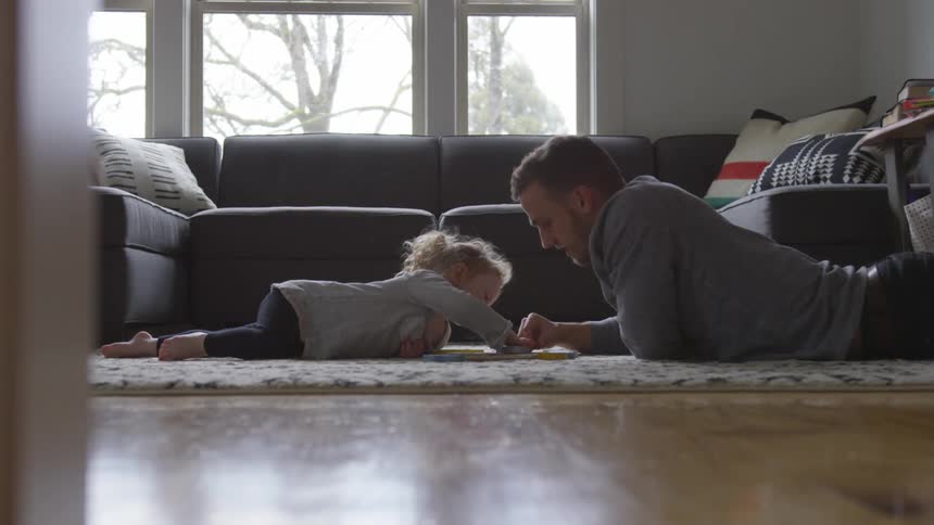 A man and his daughter lay on a rug in the living room working on a puzzle together in front of the couch