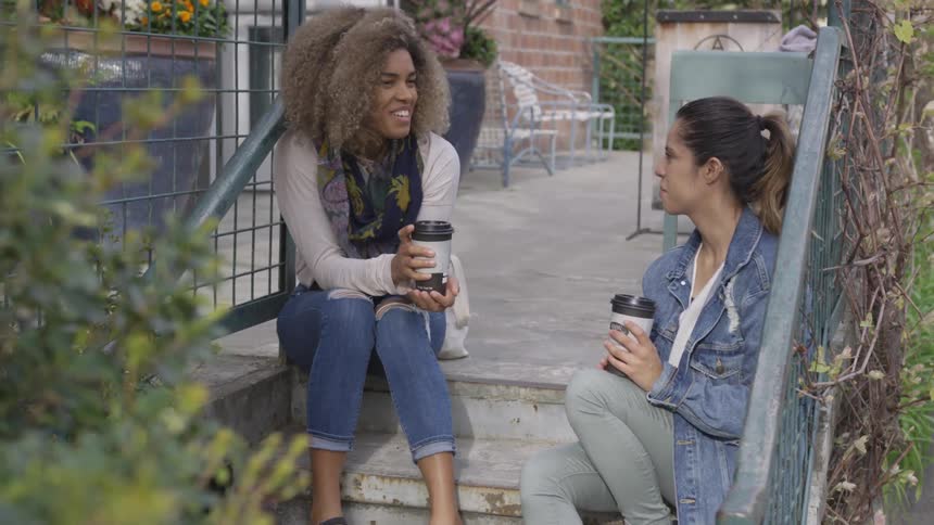 Two women talk and sit on stairs with coffee