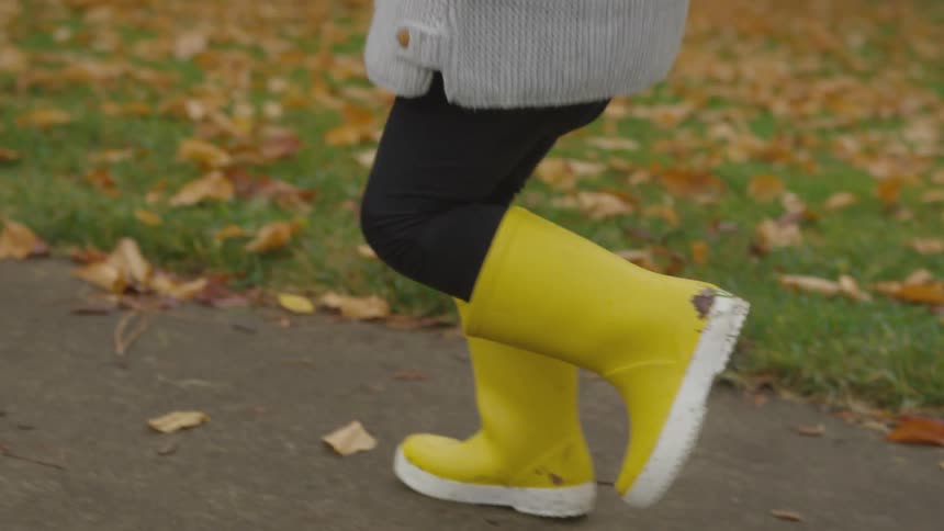 A young child runs on a sidewalk while wearing yellow rain boots on an autumn day