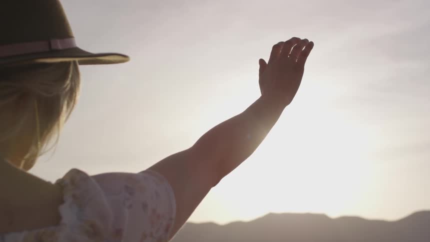 Woman wearing hat facing into sun raises hand