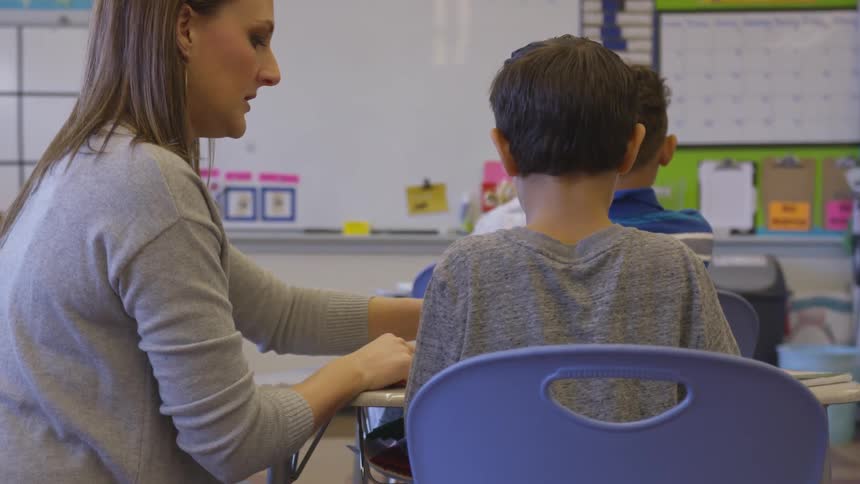 Woman helps boy while in classroom with other students