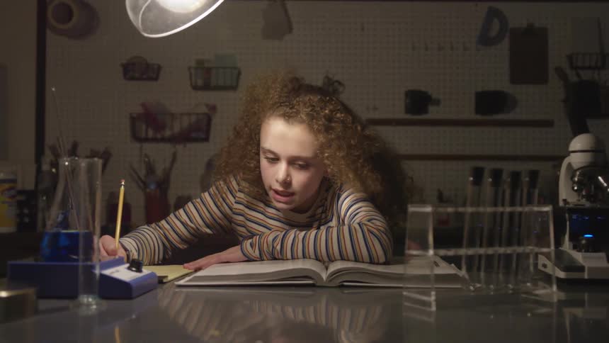 Girl sits at a table and does homework under the light of a lamp
