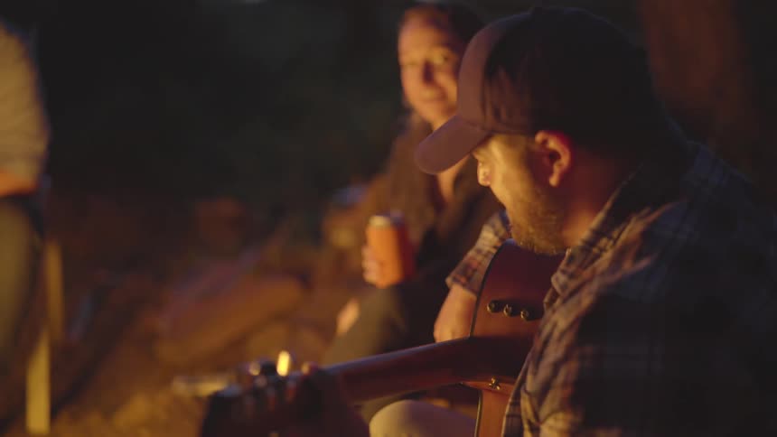 Man plays guitar with woman and others watching near campfire at night