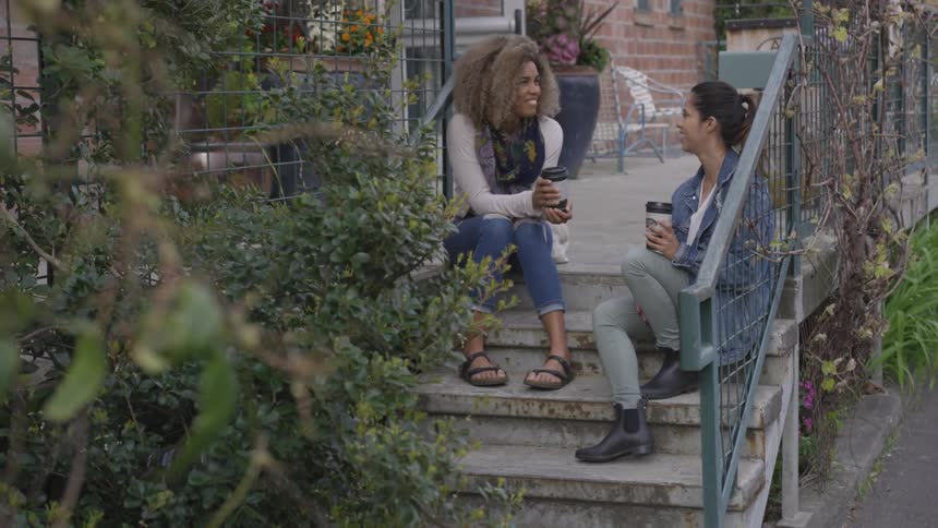 Two women sit on steps talking and holding coffee cups