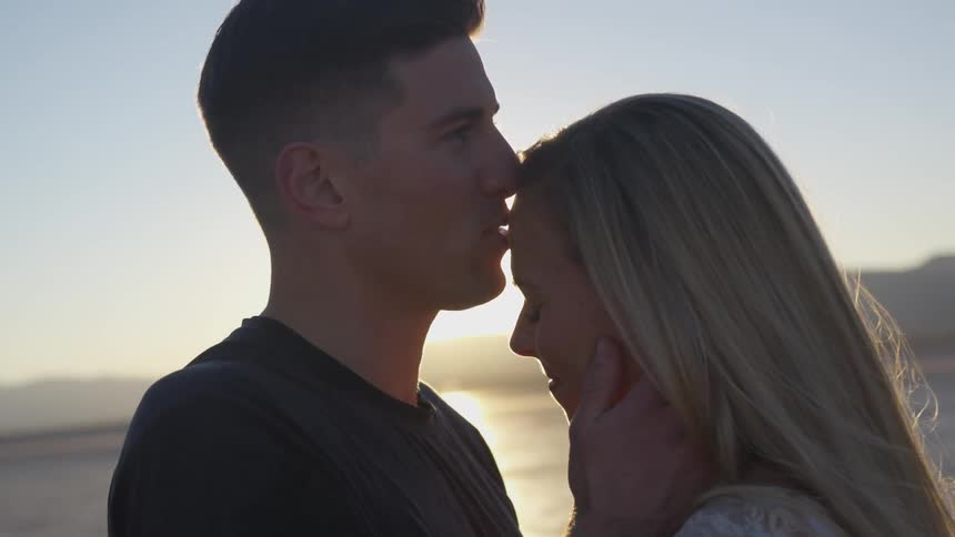A man holds a woman's face and kisses her forehead at sunset