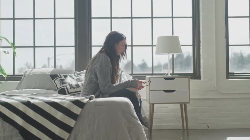A woman holds a pair of glasses while sitting on a bed