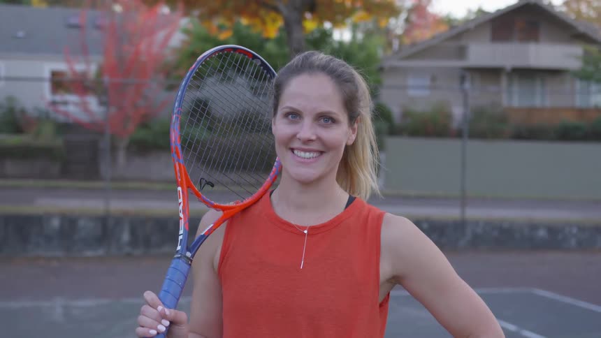 A smiling woman holds her tennis racket up against her right shoulder
