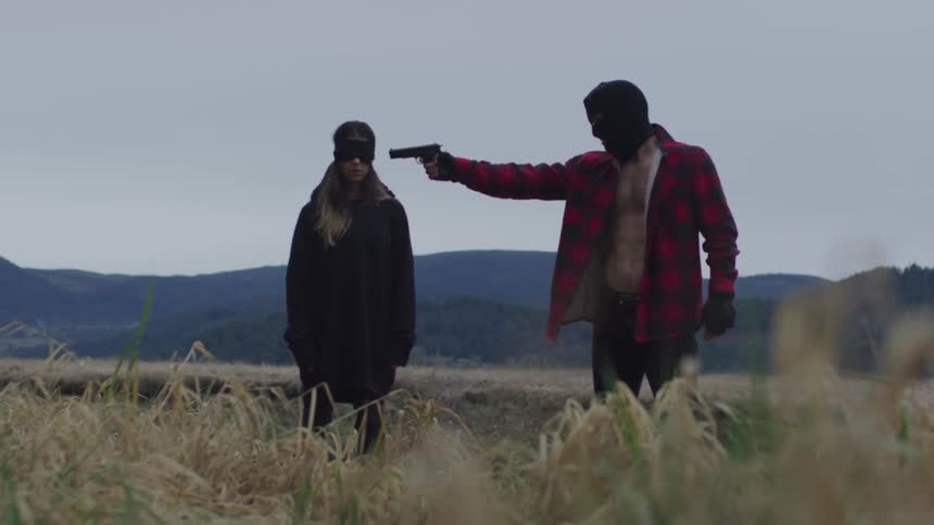 A man wearing a ski mask points a gun at a blindfolded woman outside